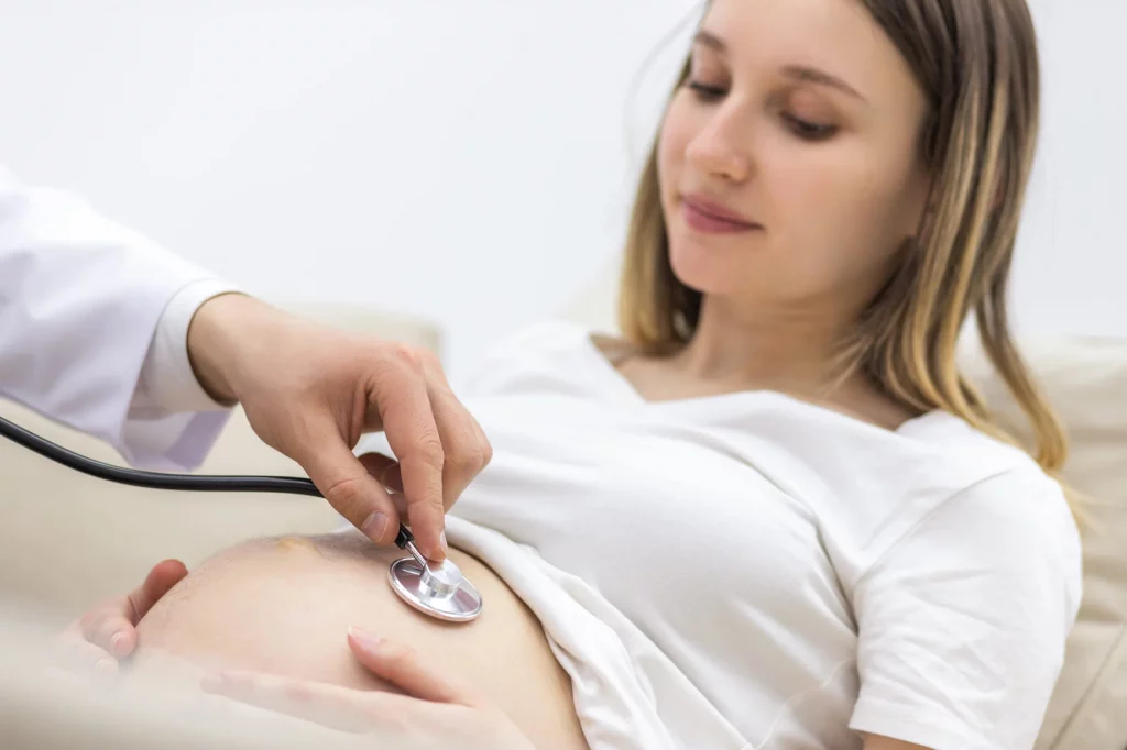 Ein Arzt hört mit einem Stethoskop die Herztöne eines Fötus am Bauch einer schwangeren Frau